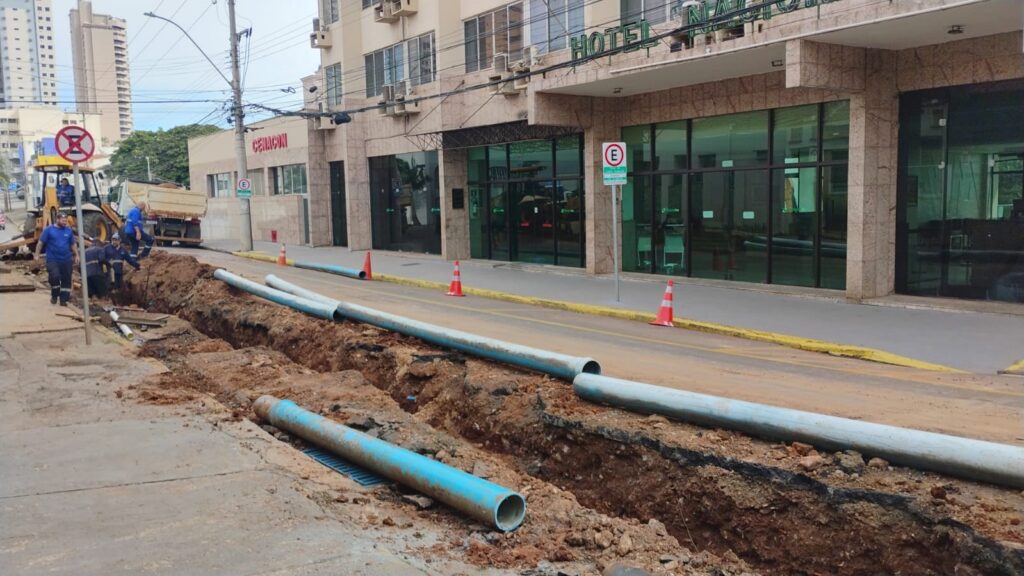 A imagem mostra uma cena de construção ao ar livre em uma rua urbana durante o dia. Há uma vala longa e estreita aberta no asfalto ao lado da calçada. Vários tubos de PVC azul claro estão dispostos ao longo da vala, alguns dentro dela e outros sobre o chão ao lado. Ao fundo, há um prédio de vários andares com a inscrição "HOTEL NACIONAL" em letras grandes, além de outro edifício com a placa "CENACON". Um grupo de trabalhadores da construção, vestindo camisetas azuis e equipamentos de segurança, está reunido próximo a uma máquina pesada (retroescavadeira) posicionada à esquerda da imagem. Vários cones de trânsito laranja delimitam a área para alertar os motoristas. O céu está nublado, e a cena transmite a ideia de manutenção ou instalação de infraestrutura urbana, possivelmente relacionada à rede de água, esgoto ou cabos subterrâneos. A perspectiva da foto é tirada da calçada, olhando ao longo da vala e dos tubos.