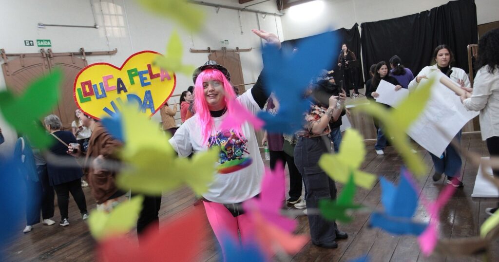 Esta imagem captura um momento de descontração e celebração em um ambiente interno, possivelmente um salão ou centro comunitário. * No centro, uma pessoa com uma peruca rosa brilhante e um chapéu está sorrindo e acenando, vestindo uma camiseta com personagens do filme "Divertidamente". Ela segura um grande cartaz em formato de coração amarelo com a frase "LOUCA PELA VIDA" escrita em letras coloridas. * Em primeiro plano, há diversos recortes de papel em formato de pássaros, nas cores verde, azul e rosa, suspensos ou espalhados, criando um efeito de desfoque que dá profundidade à cena. * Ao redor, outras pessoas participam do evento; algumas seguram cartazes brancos, enquanto outras conversam ou caminham pelo espaço. Ao fundo, percebe-se um pequeno palco onde uma pessoa parece falar ao microfone, sugerindo a realização de uma palestra ou atividade em grupo. O ambiente possui piso de madeira e portas de estilo rústico com placas de "SAÍDA".
