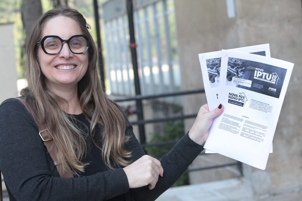 A imagem apresenta uma mulher sorridente, em plano médio, segurando documentos oficiais. Ela possui cabelos longos e castanhos, veste uma blusa preta e usa óculos de armação preta e arredondada. Na mão esquerda, ela segura dois exemplares de um documento informativo da Prefeitura de Piracicaba, referente ao IPTU 2026. O documento destaca o slogan "Nova PGV, Novo IPTU" e explica que o imposto é destinado a áreas essenciais como saúde, segurança e educação. O fundo da imagem é desfocado, mostrando uma estrutura arquitetônica com grades metálicas e uma parede de concreto, sugerindo um ambiente externo ou uma área de acesso a um prédio público. A iluminação é clara e natural, realçando a expressão positiva da pessoa.