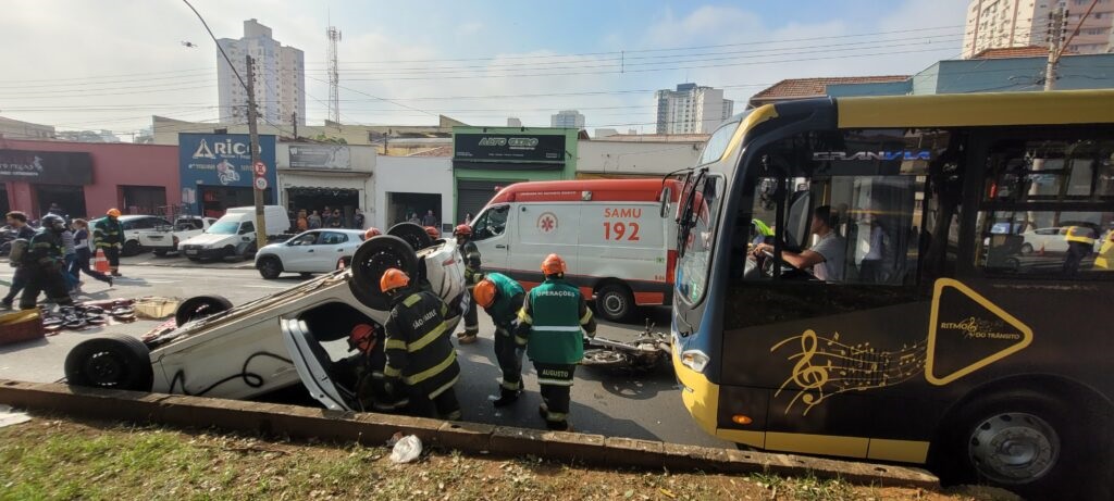 A imagem mostra a cena de um acidente de trânsito em uma área urbana durante o dia. No centro da imagem, um carro branco está tombado de lado sobre um canteiro central com grama. Vários bombeiros, usando uniformes pretos e capacetes laranja, estão ao redor do veículo, alguns aparentemente verificando o interior do carro. Ao lado, há uma ambulância do SAMU (Serviço de Atendimento Móvel de Urgência) com o número 192 visível, indicando que equipes de emergência estão atendendo a ocorrência. À direita da imagem, um ônibus preto com detalhes amarelos está parado, e o motorista está visível pela janela. O ônibus tem uma decoração musical com uma clave de sol e partituras, além da frase "RITMO DO TRÂNSITO". Ao fundo, é possível ver prédios residenciais e comerciais, além de outras pessoas e veículos que parecem estar parados ou observando a cena. A imagem transmite um momento de socorro e atendimento emergencial em uma via movimentada, com a presença de equipes profissionais e veículos de resgate. A iluminação indica que é um dia ensolarado.