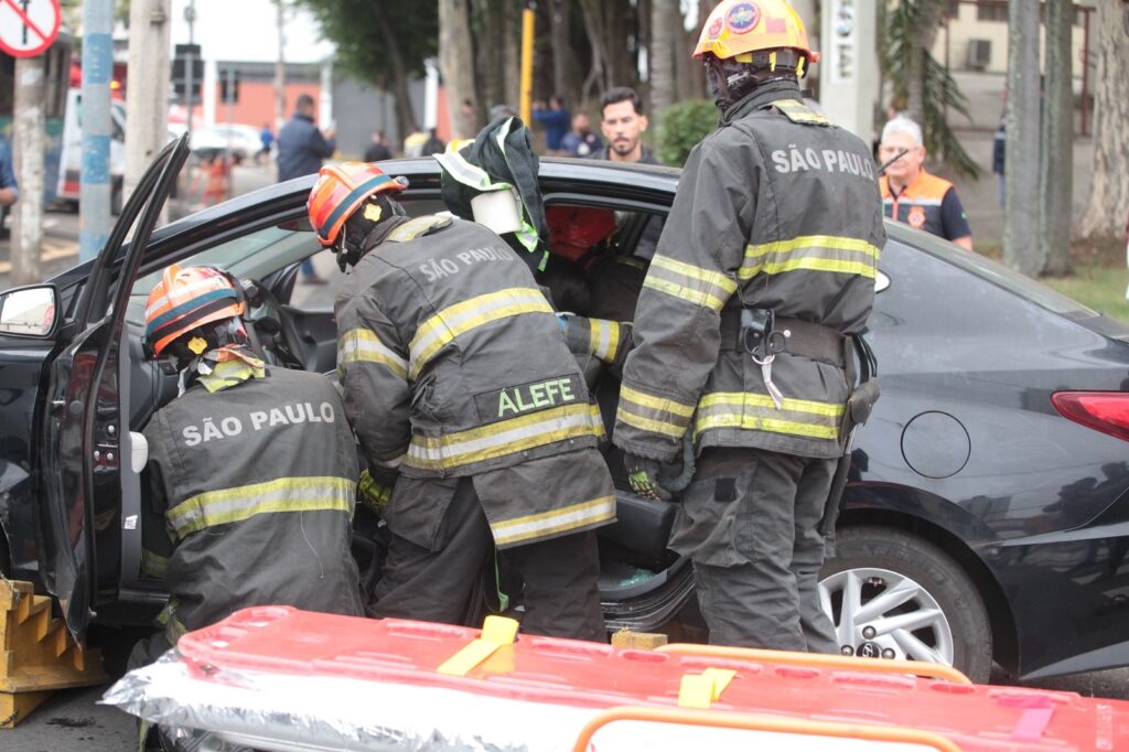 A imagem mostra uma cena de resgate em uma rua, onde três bombeiros estão auxiliando uma pessoa dentro de um carro preto, possivelmente após um acidente. Os bombeiros vestem uniformes de proteção cinza escuro com faixas refletivas amarelas e capacetes laranja. Nas costas dos uniformes está escrito "SÃO PAULO" e em um deles o nome "ALEFE". Eles estão concentrados na porta do motorista, que está aberta, tentando ajudar quem está dentro do veículo. Na frente do carro, há uma prancha de resgate vermelha, usada para imobilizar e transportar vítimas com segurança. Ao fundo, algumas pessoas observam a situação, incluindo um homem com colete laranja, possivelmente outro profissional de emergência ou segurança. A cena transmite a urgência e o cuidado da equipe de resgate no atendimento à vítima.