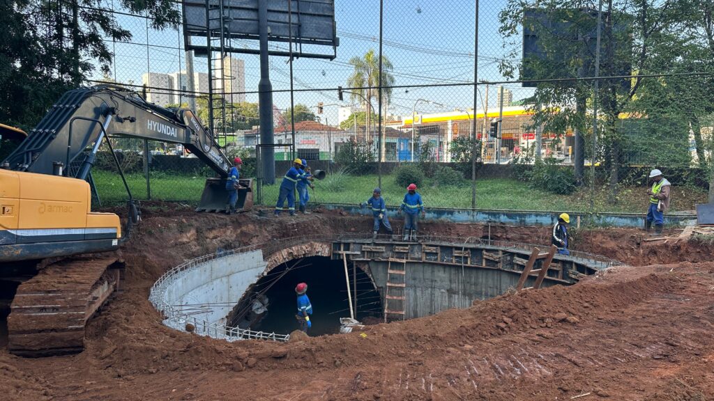 A imagem mostra um canteiro de obras com uma grande escavação em formato semi-circular, provavelmente para a construção de uma estrutura subterrânea, como um túnel ou uma galeria. As paredes da escavação são reforçadas com concreto armado e há uma abertura em arco ao fundo. À esquerda, há uma escavadeira amarela da marca Hyundai, com a inscrição "armac" em sua lateral. No local, há vários trabalhadores usando capacetes de cores diferentes (vermelho, azul, amarelo e branco) e roupas de segurança, alguns estão na borda da escavação e outros dentro dela. O solo é de terra vermelha, típica de áreas de construção. Ao fundo, é possível ver uma cerca de arame e, mais distante, um posto de gasolina da Shell. O ambiente está claro, indicando que a foto foi tirada durante o dia. No canto superior direito da escavadeira, há a inscrição "VERDEBIANCO ENGENHARIA", indicando a empresa responsável pela obra.