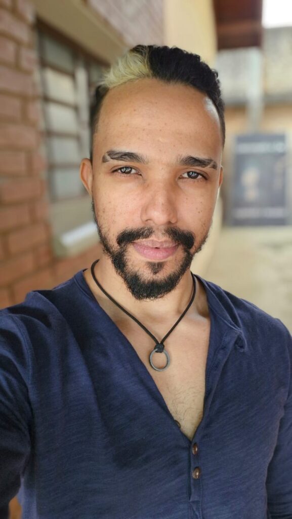 A imagem mostra um retrato em plano médio de um homem de pele clara, olhando diretamente para a câmera com uma expressão serena. Ele tem cabelos curtos, com uma parte tingida de loiro platinado e o restante escuro. Suas sobrancelhas possuem um detalhe estilizado com um pequeno corte (risco) em cada uma. Ele usa uma barba curta e bem aparada que contorna o rosto e o queixo. O homem veste uma camiseta de gola henley azul-escura com botões aparentes e utiliza um colar simples com um pingente circular metálico em um cordão preto. O fundo está levemente desfocado, sugerindo um ambiente externo com uma parede de tijolos à esquerda e uma área clara e neutra à direita, criando um contraste suave que destaca o rosto do indivíduo. A iluminação é natural e difusa, ressaltando os detalhes faciais.