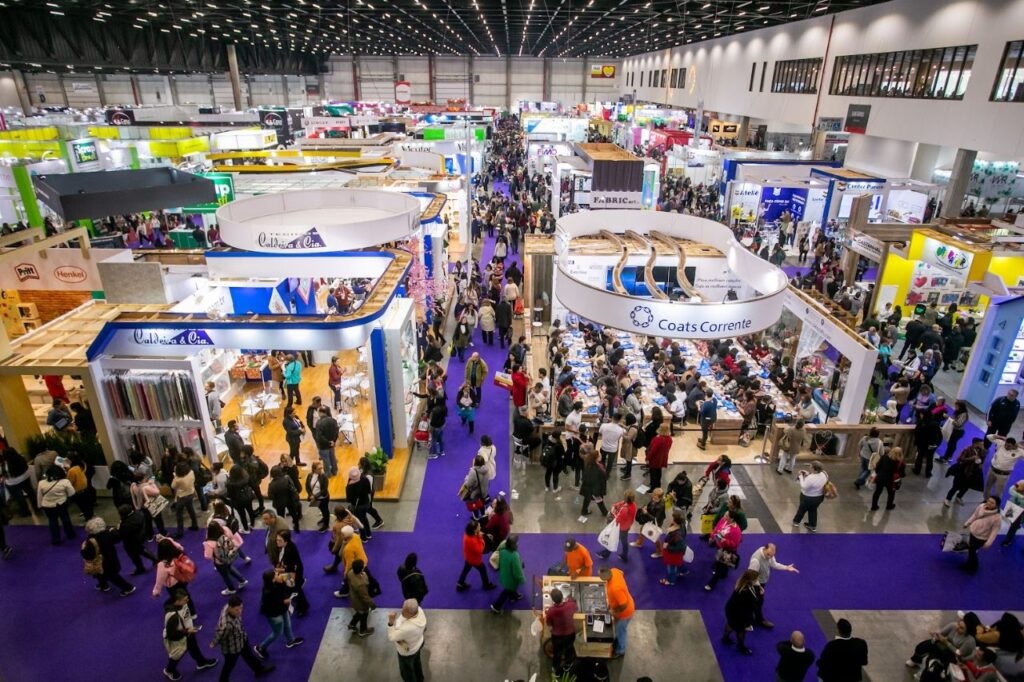 A imagem mostra uma feira ou convenção movimentada, vista de cima, com muitas pessoas caminhando por corredores cobertos por um tapete roxo vibrante. O espaço é amplo, com um teto alto e estrutura metálica aparente. Diversos estandes estão organizados ao longo do local, alguns com placas circulares suspensas indicando nomes como "Caldeira & Cia", "Coats Corrente", "Pritt", "Henkel" e "FABRIC art". A iluminação é forte e uniforme, típica de ambientes internos de eventos. A multidão é diversificada, com pessoas de diferentes idades interagindo ou circulando pelos corredores. A atmosfera parece animada e agitada, típica de uma feira comercial ou exposição de produtos.