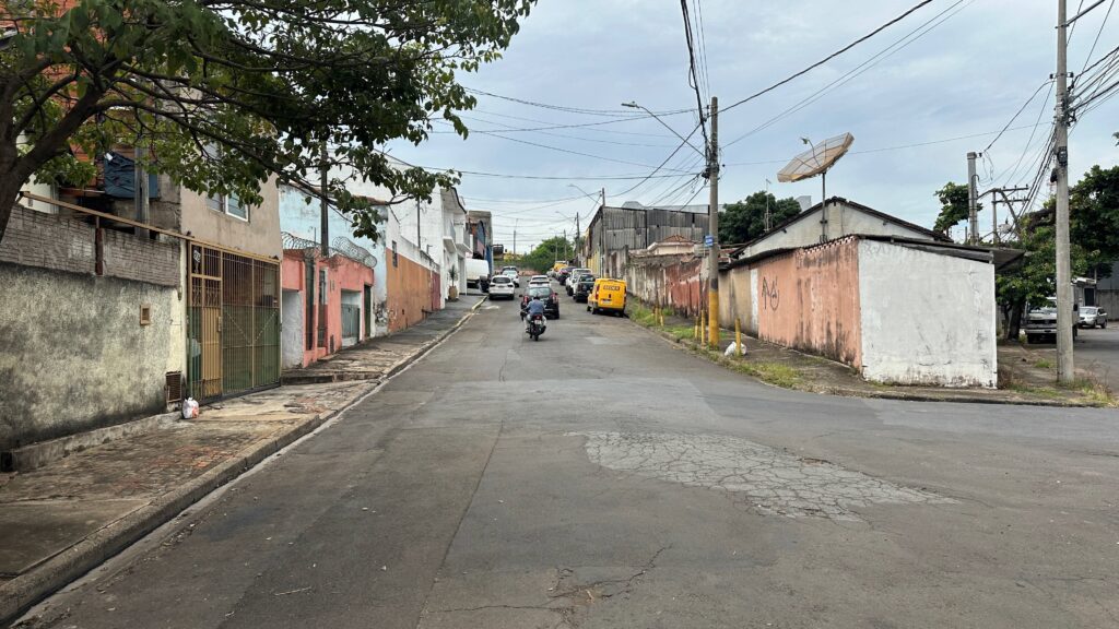 A imagem mostra uma rua pavimentada em uma área residencial ou semi-industrial, provavelmente em uma cidade do Brasil, dado o veículo SEDEX à vista. A rua está um pouco desgastada, com algumas rachaduras e remendos visíveis. Há uma motocicleta preta à frente, seguindo pelo meio da via, e mais adiante uma fila de veículos parados, incluindo uma van amarela do serviço de entregas SEDEX. As construções ao longo da rua são simples, de um ou dois andares, com paredes de cores claras e um pouco desgastadas, algumas protegidas por grades e arame farpado no topo. Há também um muro comprido à direita, com grafites e marcas de desgaste. No topo da imagem, vários fios de energia elétrica cruzam o céu nublado, que está cinza e sem sol. No geral, a cena transmite a sensação de um bairro comum, com uma atmosfera calma e cotidiana, em um dia nublado.