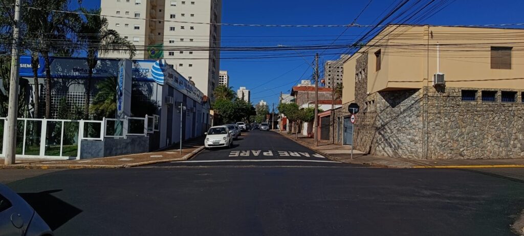 A imagem mostra uma rua urbana pavimentada, sob um céu azul limpo e ensolarado. No centro da via, há uma sinalização horizontal pintada no asfalto com a palavra "PARE" escrita duas vezes, indicando obrigatoriedade de parada para os veículos. À esquerda, observa-se uma edificação comercial moderna com grades brancas e uma placa da marca Siemens, além de um prédio residencial alto ao fundo. À direita, há uma construção com fachada revestida em pedras, típica de arquitetura local. Alguns carros estão estacionados ou em movimento ao longo da rua, que segue em direção a um horizonte onde se vislumbram outros edifícios. A cena é atravessada por uma densa rede de fios elétricos e de comunicação, caracterizando o cenário urbano de uma cidade brasileira.