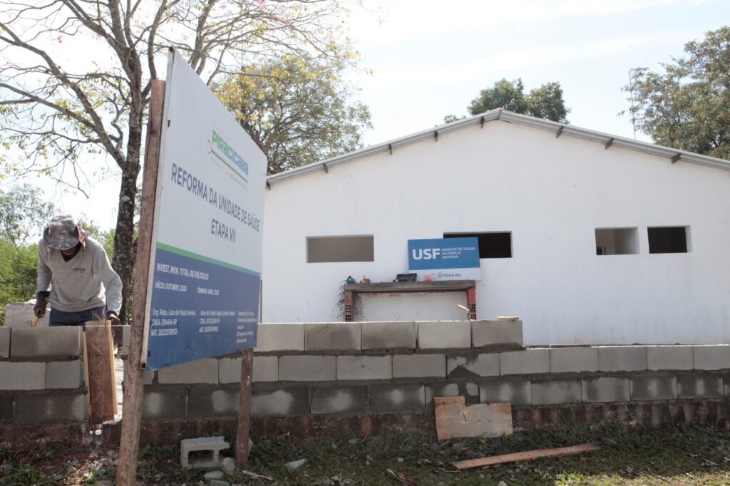 A imagem mostra uma obra em andamento, que parece ser a reforma da Unidade de Saúde da Família Santana, lá em Piracicaba, Brasil. Aqui estão os detalhes que aparecem: A Placa de Obra: Logo à frente, tem uma placa informando que se trata da "Reforma da Unidade de Saúde - Etapa VII". O investimento é de R$ 659.000,00 e o cronograma previsto vai de outubro de 2025 até abril de 2026. O Trabalho: Dá para ver um operário ali do lado esquerdo, trabalhando na estrutura de alvenaria, usando equipamento de proteção. O Local: Ao fundo, o prédio da própria unidade de saúde, pintado de branco, com a placa azul indicando o nome "USF - Unidade de Saúde da Família Santana". O ambiente parece ser bem arborizado e é um dia ensolarado.