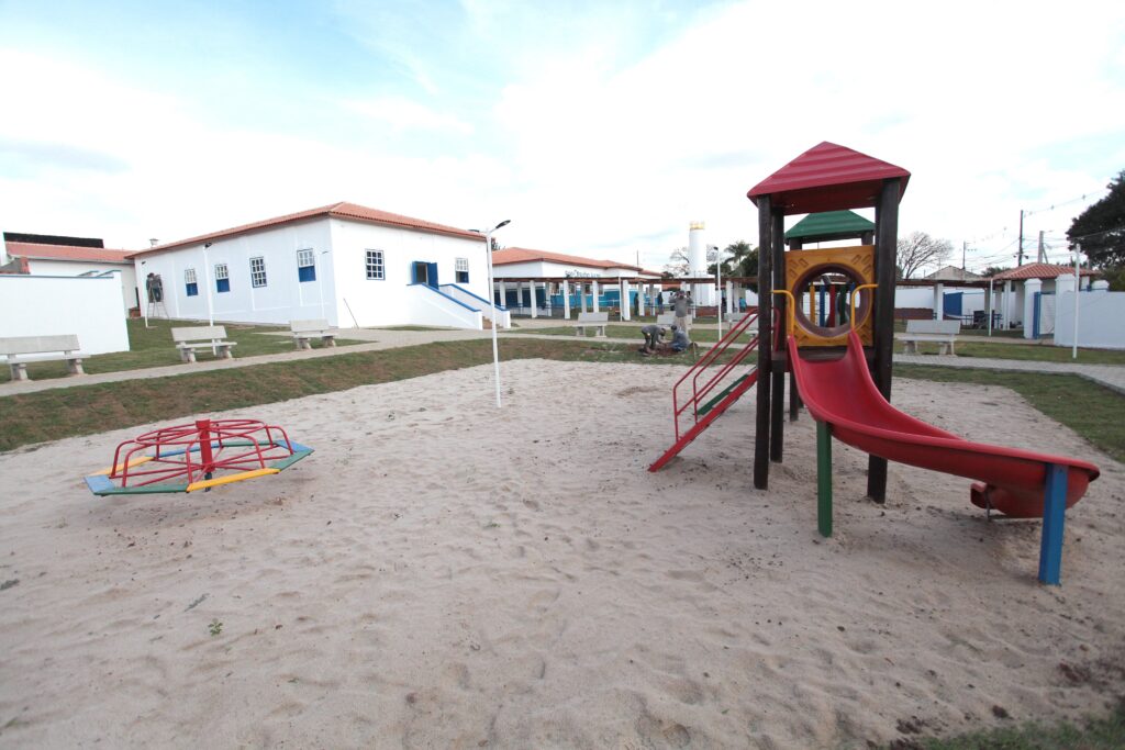 O que mostra: uma área externa de recreação infantil — um parquinho com tanque de areia. À esquerda há um brinquedo giratório (carrossel) e à direita uma estrutura de playground com escorregador e túnel. Ao fundo aparece um edifício de alvenaria com telhado de telhas cerâmicas; também há bancos de concreto e algumas pessoas ao fundo fazendo trabalhos no gramado/solo (parece jardinagem ou manutenção). Texto legível: na fachada do prédio, lê-se parcialmente algo como "SUE OTSUBO KAZU" (ou texto muito próximo disso). Formato do arquivo: imagem JPEG (.jpg). Tamanho aproximado: 7,81 MB. Iluminação e ambiente: luz suave que sugere início de manhã ou fim de tarde, céu levemente nublado. O local aparenta estar em fase de manutenção ou organização do espaço exterior.