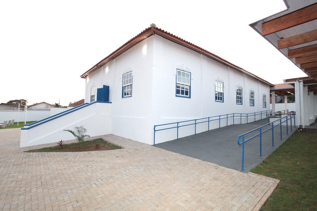 A imagem exibe uma construção térrea de arquitetura colonial com paredes brancas e janelas emolduradas em azul, situada em um ambiente externo bem cuidado. O edifício possui um telhado de telhas cerâmicas e uma estrutura de rampa de acesso para pessoas com mobilidade reduzida, equipada com corrimãos azuis, que percorre a lateral da fachada. No primeiro plano, há um pátio pavimentado com blocos de concreto intertravados, contrastando com áreas gramadas e um pequeno jardim com plantas jovens. À direita, observa-se uma estrutura de cobertura moderna em madeira, sugerindo uma área de convivência ou circulação coberta. O céu está claro e o ambiente transmite uma sensação de organização e acessibilidade.