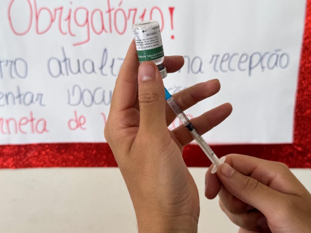 A imagem mostra um close-up das mãos de uma pessoa preparando uma vacina. A pessoa está segurando uma seringa e um frasco de vacina, enquanto parece estar puxando o líquido do frasco para a seringa. O frasco é pequeno, de vidro transparente, com uma etiqueta branca e verde contendo informações sobre a vacina, escrita em português. A seringa é transparente, com marcações visíveis, e possui uma tampa azul na agulha. Ao fundo, há um texto manuscrito em português, parcialmente legível, que menciona termos relacionados à vacinação, como "Obrigatório", "vacina", "recepção" e "uso único". A imagem é iluminada de forma clara, destacando os detalhes do frasco e da seringa. A composição transmite a ideia de um momento de preparação para uma aplicação vacinal, ressaltando a importância da vacinação e cuidados médicos.