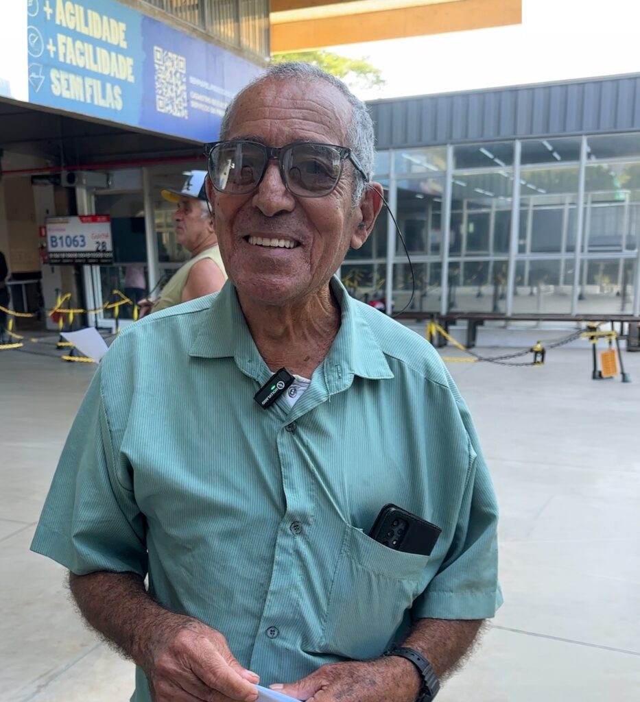 A imagem mostra um senhor de pele morena e cabelos grisalhos, sorrindo para a câmera em um ambiente que parece ser um terminal de transporte ou centro de atendimento. Ele veste uma camisa social de mangas curtas na cor verde-água, com um bolso no peito onde um celular está guardado. Em sua gola, está preso um pequeno microfone de lapela sem fio. O senhor usa óculos de grau com armação escura, que possuem uma cordinha pendurada. Ao fundo, desfocado, é possível ver outro homem de boné, além de uma estrutura metálica com sinalizações, incluindo um painel que indica "B1063" e "28", além de uma placa azul com os dizeres "+ AGILIDADE + FACILIDADE SEM FILAS" e um código QR. O chão é de concreto claro e há correntes de sinalização amarela e preta delimitando o espaço.