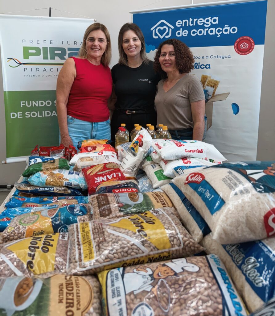 A imagem mostra três mulheres sorridentes posando atrás de uma mesa repleta de alimentos básicos, como arroz, feijão, macarrão, óleo de cozinha e outros itens de marcas variadas. Elas estão em um ambiente interno, possivelmente em um evento de doação ou assistência social. Ao fundo, há dois banners: um deles com a identificação "PREFEITURA PIRACICABA FUNDO SOCIAL DE SOLIDARIEDADE" e outro com o logo "entrega de coração". A cena transmite uma atmosfera de solidariedade e apoio comunitário, sugerindo que os alimentos estão sendo distribuídos para ajudar famílias ou pessoas em situação de vulnerabilidade.
