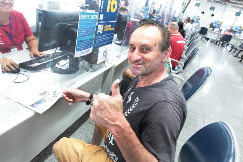 A imagem mostra um homem de meia-idade, com barba por fazer e vestindo uma camiseta preta e bermuda bege, sentado em um escritório de atendimento ao público. Ele está sorrindo e fazendo um sinal de positivo para a câmera com a mão direita, enquanto segura um documento com a esquerda. À sua frente, uma atendente vestindo uma blusa vermelha opera um computador. Entre eles, há uma divisória de acrílico transparente. No balcão, é possível ver um cartaz informativo sobre o "REFIS 2026" de Piracicaba, contendo detalhes sobre descontos para quitação de dívidas. O ambiente é um escritório amplo e bem iluminado, com várias estações de atendimento numeradas, cadeiras vazias e outras pessoas ao fundo aguardando ou sendo atendidas, sugerindo um local de atendimento municipal ou financeiro.