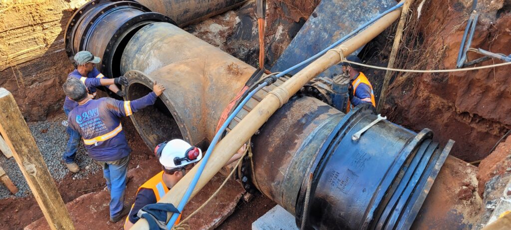 A imagem mostra uma cena de construção dentro de uma vala profunda, onde três trabalhadores estão instalando ou reparando uma grande tubulação industrial. Um dos trabalhadores, vestido com uma camiseta azul que tem o logo da empresa "AMJ" e vários números de telefone nas costas, está à esquerda, segurando a flange de uma seção de tubo em forma de "T". Outro trabalhador, usando capacete branco com protetores auriculares vermelhos e colete laranja refletivo, está no centro inferior da vala, manipulando as conexões da tubulação. Um terceiro trabalhador, também com colete laranja e boné, está à direita, próximo a uma válvula na tubulação. As tubulações são grandes, metálicas, com sinais de oxidação. A vala tem paredes de terra exposta, com raízes visíveis, e há um tubo claro que passa diagonalmente acima das tubulações principais. A iluminação é natural, e a foto foi tirada de um ângulo elevado, olhando para dentro da vala. Na tubulação maior à direita, há inscrições gravadas no metal.