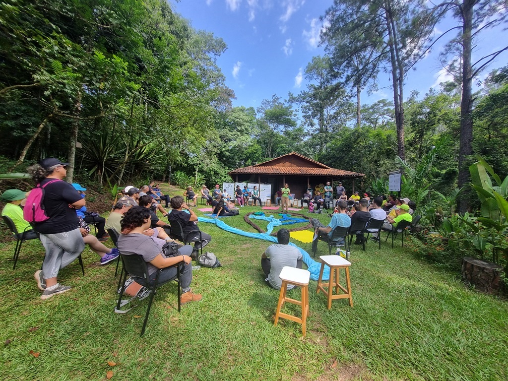 A imagem mostra um grupo de pessoas reunidas ao ar livre em uma área gramada cercada por uma floresta densa e árvores altas. * O cenário principal é um círculo formado por participantes sentados em cadeiras de plástico e bancos de madeira, voltados para uma pessoa que parece estar conduzindo uma atividade ou apresentação central. * No centro do círculo, há tecidos e materiais dispostos sobre a grama, sugerindo uma dinâmica de grupo, oficina ou atividade educativa. * Ao fundo, observa-se uma construção rústica de madeira com telhado de telhas cerâmicas, que parece ser um centro de visitantes ou um espaço de apoio. * O ambiente é iluminado pela luz do dia, com um céu azul parcialmente visível entre as copas das árvores, transmitindo uma atmosfera de harmonia, aprendizado e conexão com a natureza.