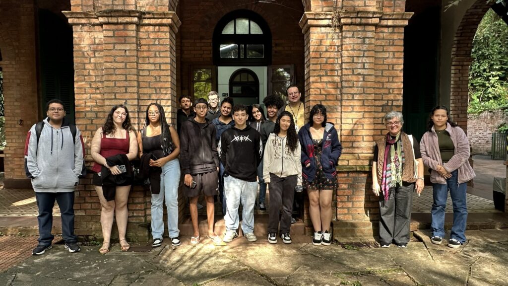 Esta imagem mostra um grupo diversificado de 15 pessoas posando para uma foto em frente a uma construção histórica de tijolos aparentes. * O grupo é composto majoritariamente por jovens adultos, com a presença de um homem mais velho ao fundo e uma mulher mais velha à direita, possivelmente professores ou orientadores. * As pessoas estão dispostas em duas fileiras, algumas em pé e outras levemente posicionadas à frente, vestindo roupas casuais como moletons, calças jeans, camisetas e vestidos. * O cenário ao fundo apresenta colunas de tijolos, arcos e uma porta de madeira, sugerindo um ambiente de visitação cultural, acadêmica ou um casarão histórico. * A iluminação é natural, com sombras projetadas no chão de pedra, indicando um dia ensolarado, e o ambiente ao redor parece ser arborizado, com vegetação visível à direita.