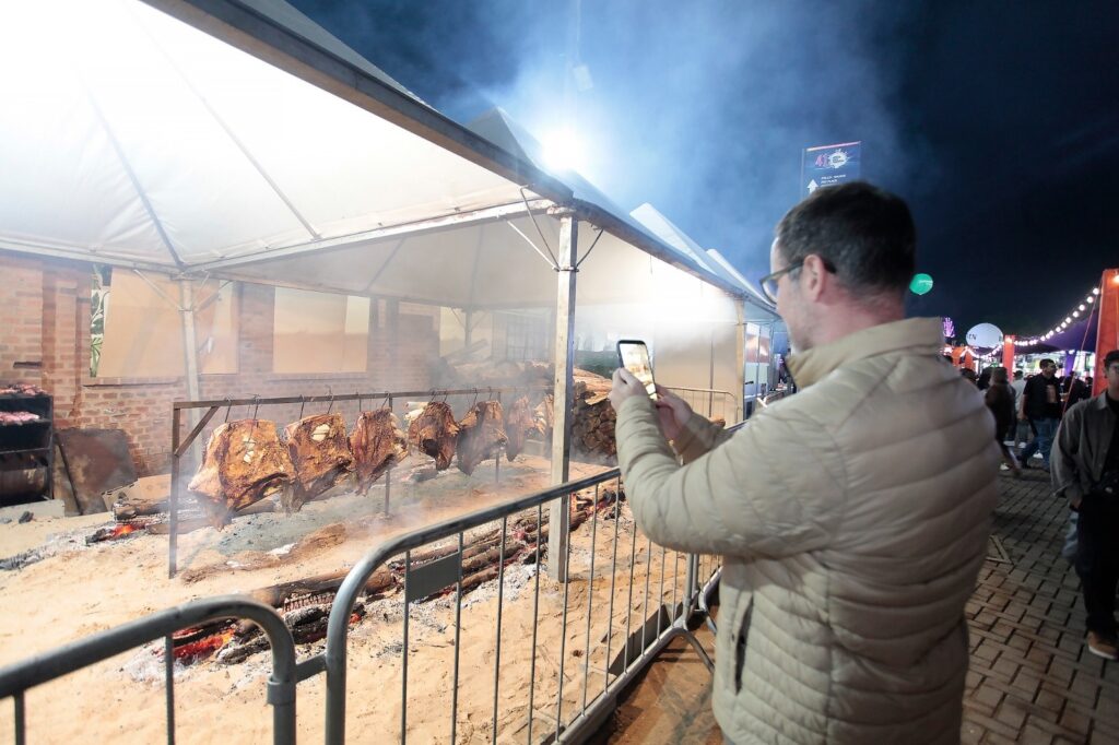 A imagem mostra uma cena noturna ao ar livre, provavelmente em uma feira ou festival gastronômico. Em primeiro plano, há um homem de cabelo castanho curto, usando óculos e uma jaqueta bege acolchoada, que está tirando uma foto com seu celular. À sua frente, dentro de uma tenda branca, grandes pedaços de carne estão sendo assados em uma estrutura metálica suspensa sobre uma fogueira com brasas e lenha. A fumaça da fogueira cria um efeito nebuloso no ambiente iluminado por luzes artificiais fortes. Ao redor da área de cozimento, há barreiras metálicas que delimitam o espaço. Ao fundo, há uma parede de tijolos, pilhas de lenha e algumas pessoas caminhando. Também é possível ver uma placa com o número "41". A cena transmite uma atmosfera típica de evento ao ar livre, com foco na preparação tradicional de carne assada.