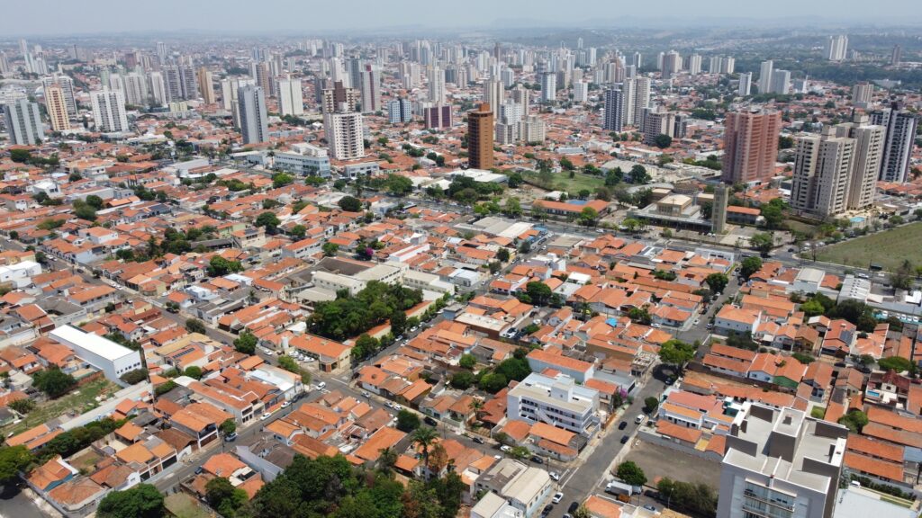 A imagem mostra uma vista aérea de uma área urbana densamente povoada, provavelmente de uma cidade brasileira, devido aos telhados de cerâmica vermelha típicos das construções residenciais. Na parte mais próxima, vemos casas baixas com telhados inclinados, ruas estreitas e algumas árvores espalhadas entre as casas. Ao fundo, a paisagem muda para prédios altos e modernos, formando um skyline urbano com muitos edifícios residenciais e comerciais. O céu está claro e o dia parece ensolarado, com luz natural que realça as cores quentes dos telhados e o cinza dos prédios. A imagem transmite a sensação de uma cidade grande, com contrastes entre áreas mais antigas e residenciais e áreas mais novas e verticalizadas.