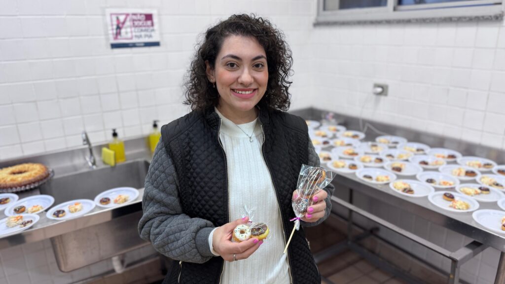 A imagem mostra uma jovem mulher de cabelos cacheados, sorrindo para a câmera, em uma cozinha industrial ou copa. Ela veste uma blusa branca de gola alta e um colete acolchoado preto. Em suas mãos, ela segura pequenos donuts decorados: na mão esquerda, dois donuts individuais dentro de uma embalagem plástica transparente, e na mão direita, um espeto com três donuts empilhados, também embalados. Ao fundo, há uma bancada de aço inoxidável repleta de pratos brancos, cada um contendo pequenos doces ou donuts, sugerindo a preparação para um evento ou festa. À esquerda, vê-se uma pia de metal com dispensadores de sabonete e, na parede, um cartaz com instruções de higiene.