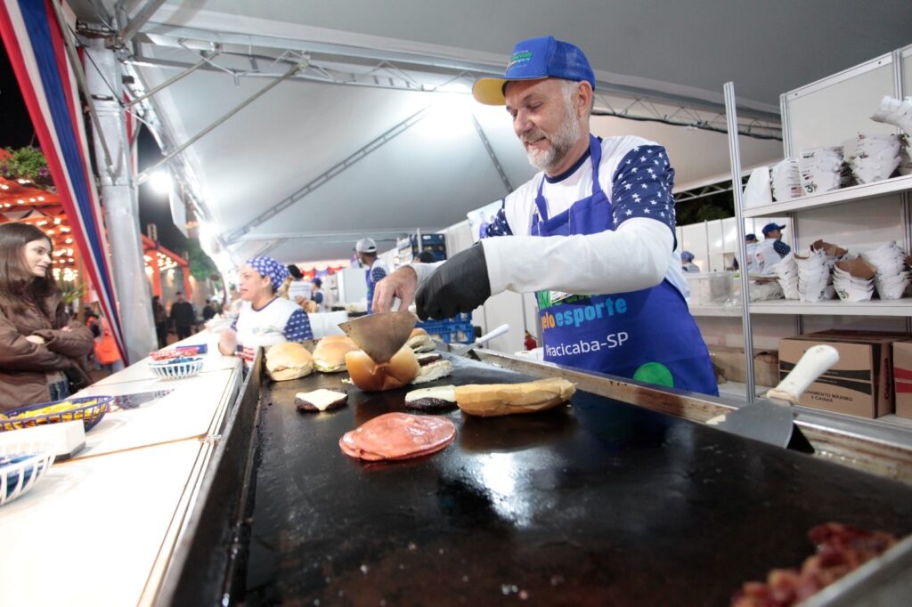 A imagem mostra uma barraca de comida em um evento ao ar livre à noite. No centro, há um homem de meia-idade com barba grisalha, usando um boné azul, uma camisa branca com estampa de estrelas nas mangas e um avental azul. Ele está concentrado em preparar hambúrgueres em uma chapa grande, usando uma espátula para virar um pão. Na chapa, há diversos hambúrgueres, fatias de queijo, presunto e pães sendo preparados. Ao lado, uma mulher com lenço azul na cabeça e camisa estampada também está trabalhando na barraca. À esquerda, uma jovem observa a preparação dos alimentos. O ambiente é iluminado por luzes artificiais típicas de tendas em eventos, e ao fundo há prateleiras com embalagens para comida. O avental do homem tem a inscrição "pelo esporte Piracicaba-SP", sugerindo que a barraca pode estar ligada a algum projeto esportivo ou comunitário da cidade de Piracicaba, São Paulo. A cena transmite a atmosfera de um festival ou feira, com foco na preparação artesanal dos alimentos e no trabalho em equipe.