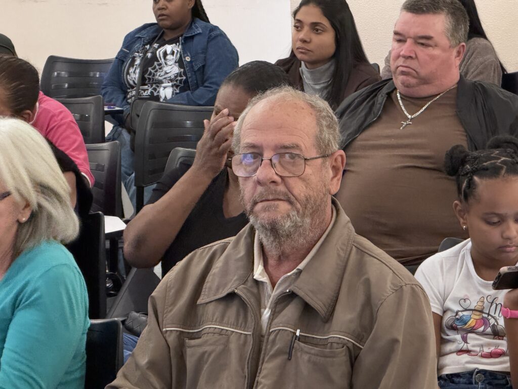 Cena interna com várias pessoas sentadas em cadeiras pretas alinhadas, parecendo assistir a uma apresentação, missa ou reunião. Foco principal: homem idoso, pele clara, cabelo grisalho curto, óculos, barba por fazer, vestindo jaqueta bege/ castanho sobre camisa clara. Ele está no centro e olha para a frente com expressão séria. Pessoas próximas: À direita do homem central: um homem mais velho e robusto, jaqueta preta, camiseta marrom e um colar com pingente de cruz. Ainda à direita, em primeiro plano: uma jovem com cabelo preso em dois coques, camiseta branca com estampa de unicórnio, segurando um celular. À esquerda, em primeiro plano: parte de uma mulher com cabelos brancos curtos e blusa azul-turquesa. Atrás do homem central: uma mulher de pele morena e cabelos escuros, com a mão próxima ao rosto (aparenta estar pensativa ou cobrindo parte do rosto). Canto superior esquerdo: mulher de jaqueta jeans sobre blusa estampada; mais ao centro, outra mulher de cabelos longos e gola alta. Ambiente: cadeiras plásticas pretas enfileiradas, iluminação suave; profundidade de campo que mantém o homem de jaqueta bege em foco, com as outras pessoas um pouco desfocadas.