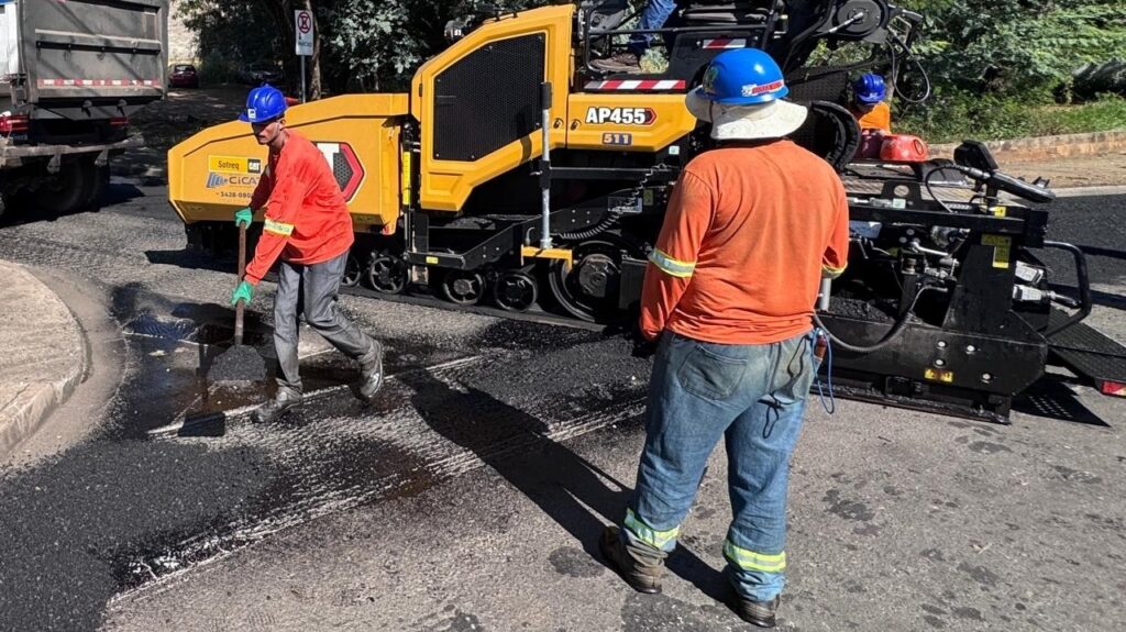 A imagem mostra uma cena de trabalho em uma obra de pavimentação asfáltica. * Em primeiro plano, um operário de costas, vestindo uma camisa laranja de manga comprida com faixas refletivas, calça jeans e um capacete azul com proteção para o pescoço, observa o processo. * À esquerda, outro operário, também usando camisa laranja, calça cinza, luvas e capacete azul, utiliza uma ferramenta manual para espalhar ou ajustar o asfalto recém-aplicado no solo. * Ao fundo, destaca-se uma pavimentadora de asfalto amarela da marca CAT, modelo AP455, que é o equipamento principal utilizado para distribuir e nivelar a massa asfáltica. * O ambiente é uma via pública, onde é possível notar parte de um caminhão basculante à esquerda, uma placa de sinalização de trânsito e vegetação ao fundo, indicando um dia ensolarado.