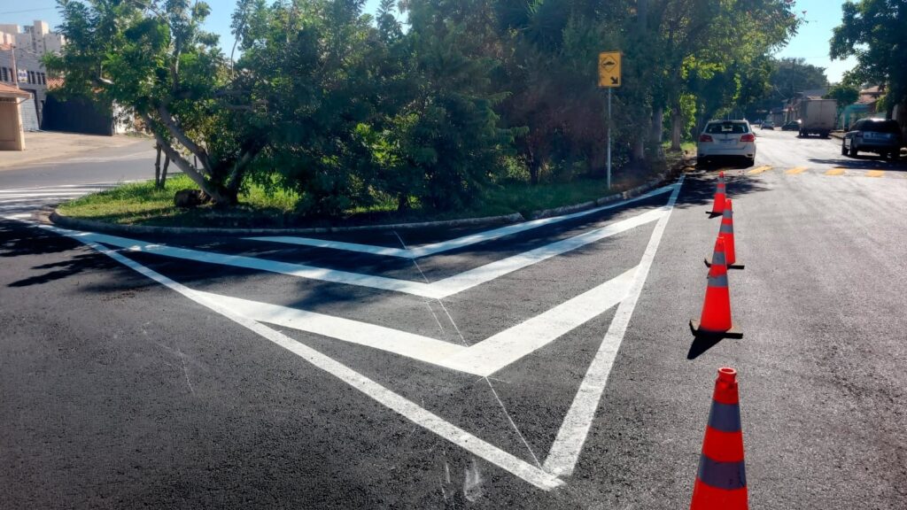 A imagem mostra uma rua pavimentada recentemente, com uma pintura de sinalização viária em formato de "V" branco no centro da pista, indicando uma área de separação ou desvio de tráfego. Ao lado direito da pintura, há quatro cones de trânsito laranja alinhados, provavelmente para orientar os motoristas a não passarem sobre a área pintada. A rua é cercada por uma calçada e, ao fundo, há uma placa amarela de sinalização que indica a presença de lombada. Também é possível ver alguns carros estacionados e árvores ao longo da via, sugerindo que é uma área residencial ou suburbana. A iluminação é natural, com luz do dia e sombras visíveis.