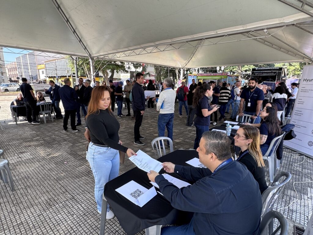 A imagem mostra uma cena de um evento ou feira de empregos ao ar livre, sob uma grande tenda branca. No centro, uma mulher de blusa preta e calça jeans entrega um documento para um homem sentado atrás de uma mesa coberta com uma toalha preta. Ao lado dele, outra mulher com óculos observa a interação. Ao redor, há diversas pessoas circulando, conversando e interagindo em outras mesas de atendimento. O ambiente parece ser uma praça ou área pública, com calçamento de pedras, árvores ao fundo e algumas barracas de comida, como uma de "Churros e Crepes". À direita, há um banner da empresa Hyundai, indicando que se trata de uma ação corporativa ou de recrutamento. A iluminação é de um dia ensolarado.