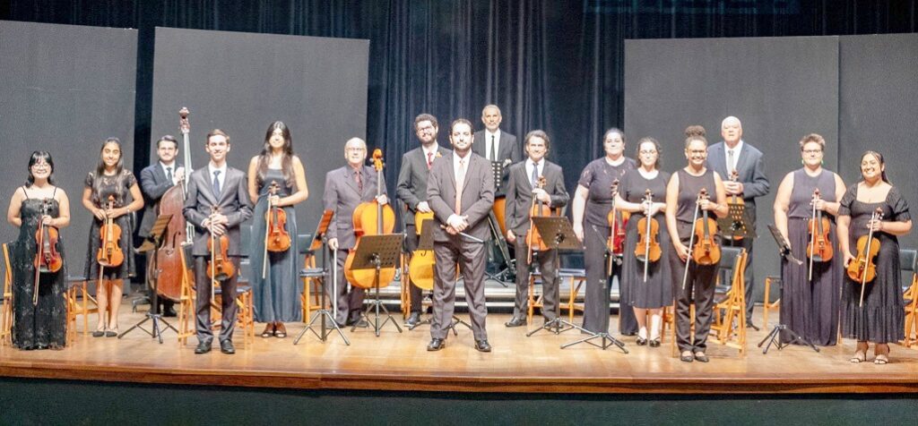 Nesta imagem, uma orquestra de câmara posa para uma foto em um palco com piso de madeira e fundo preto. O grupo é composto por músicos que vestem trajes formais (ternos ou vestidos pretos) e seguram seus instrumentos. Podemos identificar: • À esquerda, uma violinista e uma mulher segurando um violino, seguidas por um contrabaixista. • Seguindo para o centro, há um violinista, outra violinista, um violoncelista e um violinista ao lado do maestro (ou regente), que está de terno e gravata, posicionado em destaque no meio do grupo. • À direita, a orquestra continua com um músico de terno, uma musicista de preto, uma violinista, outra musicista, um homem de terno, uma violinista e, por fim, uma mulher de vestido preto à direita. Todos estão posicionados de forma organizada, muitos com seus violinos, violas ou violoncelos em mãos, preparados para uma apresentação.