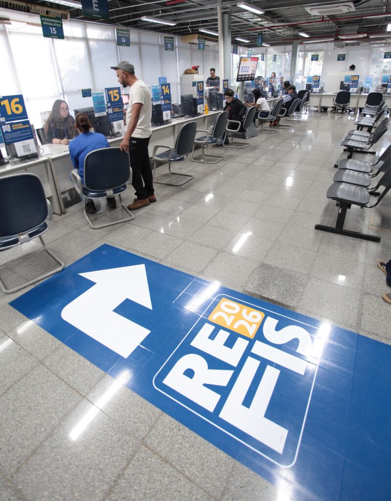 Esta foto mostra o interior de um órgão público ou escritório administrativo de atendimento ao cliente, provavelmente relacionado à área fiscal ou tributária. O espaço: É uma sala ampla com piso de granito, equipada com vários guichês de atendimento numerados (de 05 a 16 visíveis). Cada posto possui um computador e uma divisória de vidro para proteção. Há cadeiras de espera enfileiradas à direita. Atendimento: Vemos algumas pessoas sendo atendidas nos balcões, com funcionários sentados atrás das telas e cidadãos sentados ou de pé à frente. Destaque no piso: O elemento mais chamativo é um grande adesivo azul colado no chão, em primeiro plano, com uma seta branca apontando para a frente e o texto "REFIS 2026", indicando que o local está operando um programa de regularização de débitos fiscais para este ano. Atmosfera: O ambiente é funcional, organizado e bem iluminado, típico de repartições públicas ou postos de atendimento ao contribuinte.