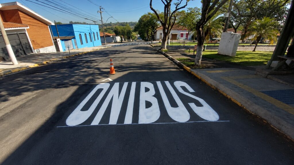 Esta foto mostra uma rua tranquila, em um dia ensolarado, com uma sinalização horizontal pintada no asfalto. A sinalização: Em destaque, lê-se a palavra "ONIBUS" escrita em letras grandes e brancas no centro da via. Logo à frente, há um pequeno cone de trânsito laranja. O entorno: À esquerda, destaca-se um prédio com fachada azul e uma parte superior em tijolos aparentes. À direita, há uma área arborizada com gramado, calçada e alguns bancos, sugerindo ser uma pequena praça ou área de convivência. Clima: O céu está limpo e a iluminação é de um dia claro, projetando sombras nítidas das árvores sobre o pavimento.