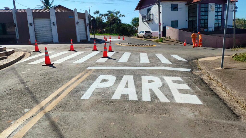 A imagem mostra uma cena de serviço público de sinalização viária em uma rua pavimentada. Em primeiro plano, destaca-se uma pintura horizontal recém-feita no asfalto com a palavra "PARE" em letras grandes brancas, seguida por uma faixa de pedestres também pintada de branco. * Vários cones de trânsito alaranjados foram dispostos ao longo da via para isolar a área e orientar o fluxo de veículos enquanto a tinta seca. * Ao fundo, à direita, dois funcionários vestindo uniformes operacionais alaranjados de alta visibilidade estão em pé, observando o trabalho. * O cenário inclui construções ao redor, como uma edificação de dois andares com fachada clara e detalhes em roxo, além de uma estrutura residencial à esquerda com portões metálicos fechados. O céu está limpo e ensolarado, indicando um dia claro.
