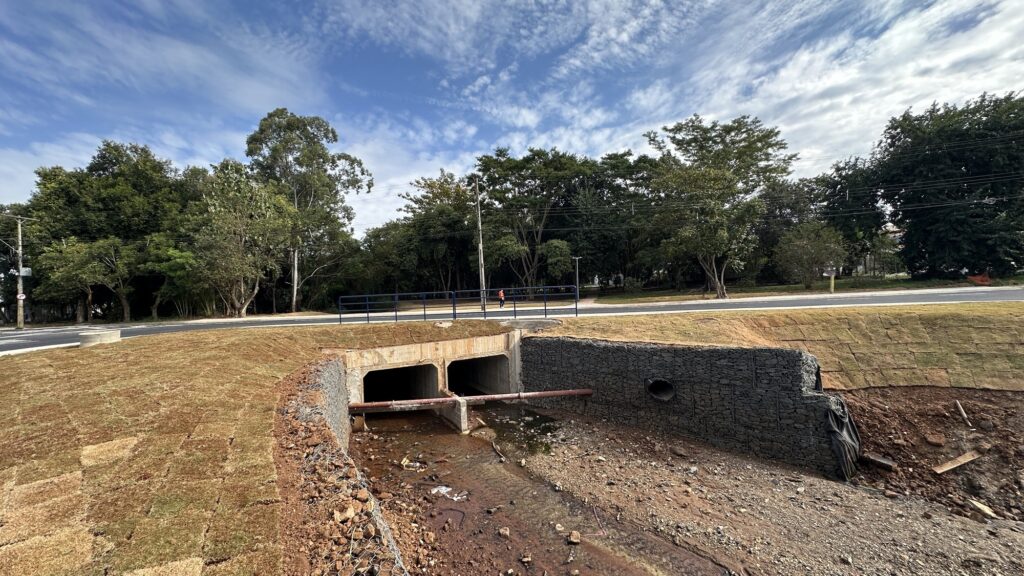 A imagem mostra uma estrutura de drenagem em uma área de construção ou engenharia civil. Trata-se de um bueiro com duas aberturas de concreto, instalado em um talude reforçado com gabiões — estruturas feitas de malha de arame preenchidas com pedras. Na frente das aberturas do bueiro, há um tubo vermelho que atravessa horizontalmente. O leito do curso d'água abaixo do bueiro é seco e coberto por pedras e detritos. As laterais do talude estão cobertas com grama recém-plantada, provavelmente para evitar erosão. Acima do bueiro, há uma estrada asfaltada com uma proteção metálica azul (guardrail). Ao fundo, vê-se uma pessoa usando uma camiseta laranja caminhando pela estrada, e uma linha de árvores verdes sob um céu azul com poucas nuvens. A iluminação indica que a foto foi tirada durante o dia, em um ambiente claro e ensolarado.