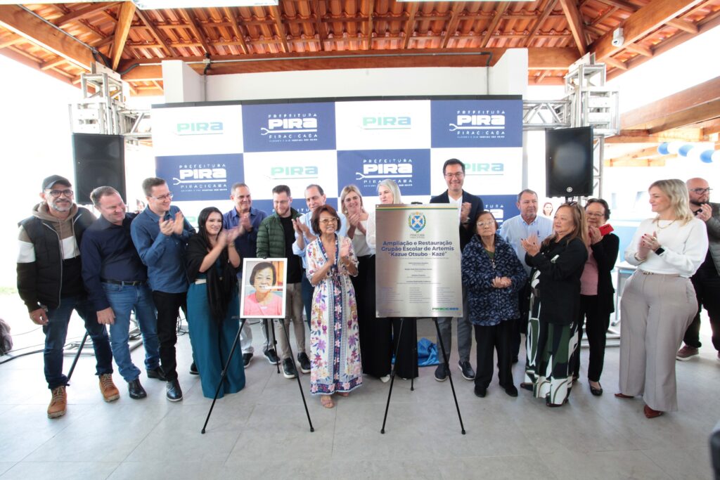 A imagem registra uma cerimônia oficial de entrega de obras da Prefeitura de Piracicaba. * O grupo está posicionado sob uma estrutura de madeira, em frente a um painel azul e branco com a marca da prefeitura. * No centro, destaca-se uma placa comemorativa intitulada "Ampliação e Restauração Grupo Escolar de Artemis 'Kazue Otsubo - Kazé'". * À esquerda da placa, uma mulher segura um retrato emoldurado da homenageada. * O grupo, composto por cerca de 15 pessoas entre homens e mulheres, está vestido com trajes casuais e formais, muitos deles batendo palmas ou sorrindo, celebrando a inauguração. * O ambiente é iluminado e sugere um evento público, com equipamentos de som visíveis nas laterais.