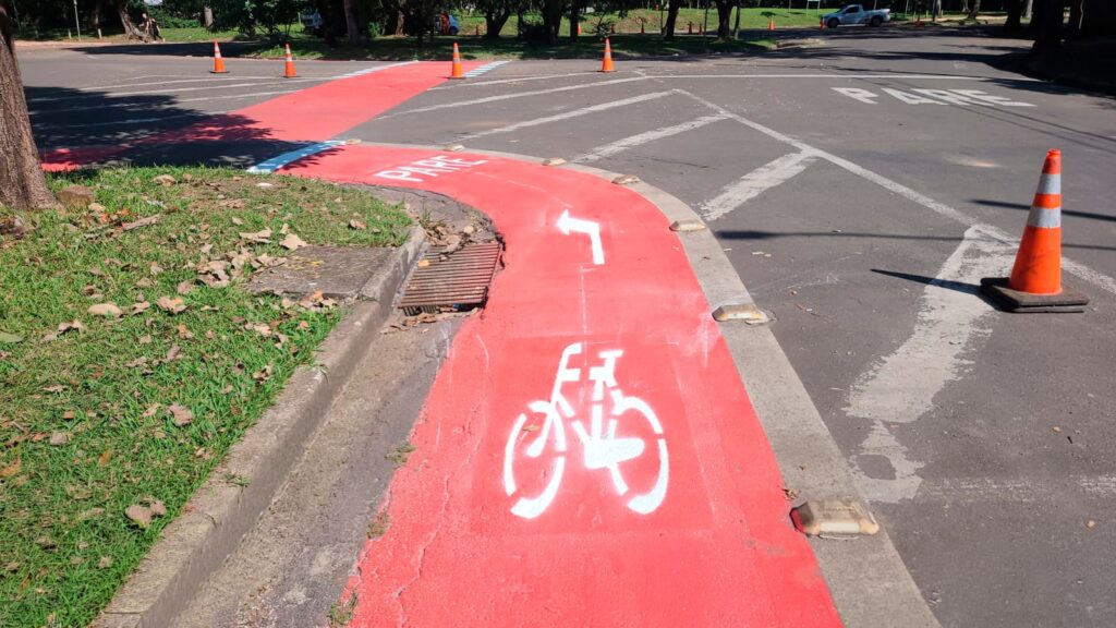 A imagem mostra uma ciclovia pintada de vermelho vibrante em uma área urbana. * O destaque central é o símbolo de uma bicicleta pintado em branco no chão, acompanhado por uma seta indicativa apontando para a esquerda, sinalizando a direção do fluxo. * A ciclovia segue o contorno de uma calçada gramada e termina em um cruzamento, onde há a palavra "PARE" escrita no pavimento, reforçando a necessidade de atenção dos ciclistas. * O ambiente inclui sinalizações de trânsito, como cones laranjas dispostos ao longo da via e marcações brancas no asfalto que delimitam a faixa de pedestres e o cruzamento. * O cenário é iluminado pela luz do dia, com sombras projetadas por árvores e pelos objetos de sinalização, sugerindo um espaço público bem estruturado para a mobilidade urbana.