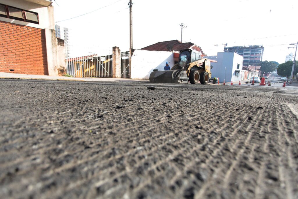 A imagem mostra uma rua em obras vista de um ângulo baixo, com o foco na superfície da estrada que foi recentemente fresada, exibindo sulcos profundos e textura áspera. No meio da imagem, há uma máquina amarela da marca Caterpillar, equipada com uma escova rotativa na frente, usada para limpar os resíduos da fresagem. Um trabalhador com capacete azul está próximo à máquina. Ao fundo, é possível ver casas e construções, além de postes de energia e cones de sinalização laranja ao longo da via, indicando que a área está em reforma. A luz do dia está difusa, sugerindo um céu nublado. A composição destaca o contraste entre a textura da superfície da estrada e os elementos da obra.