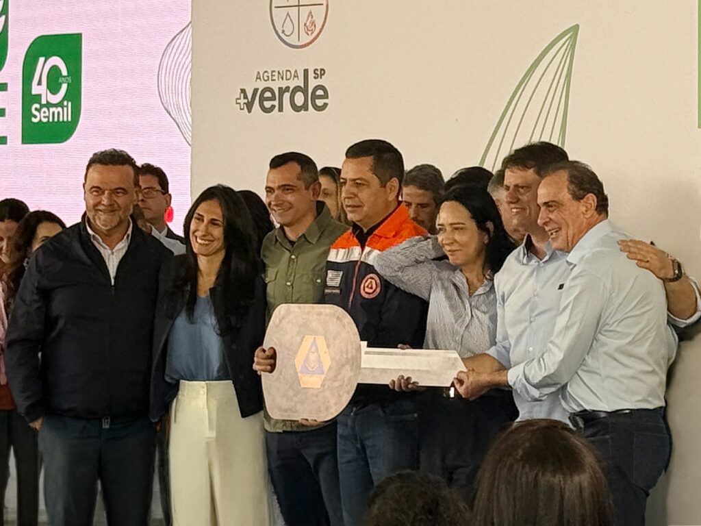 A imagem mostra um grupo de oito pessoas posando para uma foto em um ambiente interno, provavelmente durante um evento oficial relacionado à "Agenda Verde SP". No centro da imagem, um homem veste uma jaqueta laranja e azul-marinho com um logo circular e está segurando uma grande chave simbólica, que compartilha com a pessoa ao lado. As outras pessoas ao redor estão vestidas de forma casual e formal, com camisas, blusas e jaquetas. Ao fundo, há uma parede branca com o logotipo "AGENDA SP +verde" e uma inscrição que menciona "40 ANOS Semil". A iluminação é clara, típica de um ambiente fechado, e a atmosfera parece ser profissional e comemorativa. A imagem transmite um momento de celebração ou entrega simbólica dentro de um contexto institucional ou governamental.