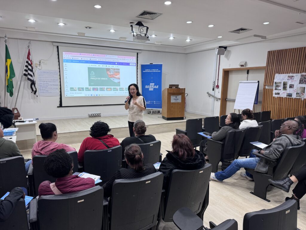 A imagem mostra uma sala de aula ou auditório onde uma mulher está ministrando uma palestra ou aula para um grupo de pessoas. Ela está em pé, próxima a um púlpito de madeira, em frente a um grande telão onde está projetada uma apresentação intitulada "AULA 6 PREÇO DE VENDA". A sala é bem iluminada, com paredes claras e um piso de madeira. À esquerda, há um cartaz na parede com o título "CONTRATO DE CONVIVÊNCIA", que lista regras como respeito, empatia, não julgamento, escuta ativa, celular no silencioso, sem bullying, sem racismo e preconceito, colaboração, paciência, pontualidade e cumprir combinado. Também há uma bandeira ou banner azul com o logo do SEBRAE e o slogan "A FORÇA DO EMPREENDEDOR BRASILEIRO", indicando que o evento é relacionado a empreendedorismo e provavelmente promovido por essa instituição. O público está sentado em cadeiras dispostas em fileiras, prestando atenção na apresentação. O ambiente é organizado e profissional, sugerindo um treinamento ou workshop voltado para empresários ou empreendedores.