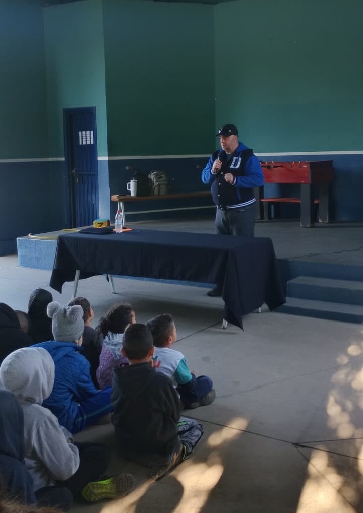 A imagem mostra um homem falando para um grupo de crianças em um ambiente interno, que parece ser uma escola ou centro comunitário. Ele está em um palco, segurando um microfone, usando uma jaqueta azul com detalhes em preto e um boné escuro. As crianças estão sentadas no chão, de costas para a câmera, prestando atenção ao que ele diz. O ambiente tem paredes azuis e uma mesa de pebolim ao fundo. A cena transmite uma atmosfera de aprendizado ou apresentação, com as crianças concentradas no orador.