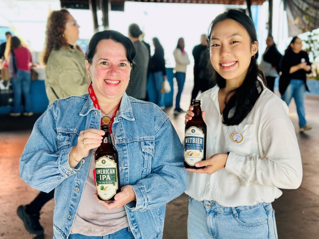A imagem mostra duas mulheres sorridentes em um ambiente interno, possivelmente uma feira ou evento, possando para uma foto enquanto seguram garrafas de cerveja artesanal da marca "Soro & Lúpulo". * A mulher à esquerda veste uma jaqueta jeans sobre uma blusa clara e usa um cordão com crachá. Ela segura uma garrafa de "American IPA" com soro de leite. * A mulher à direita veste uma camisa social branca e calça jeans, segurando uma garrafa de "Witbier", também feita com soro de leite. Ela tem um pequeno broche circular preso à camisa. * Ao fundo, nota-se um ambiente movimentado com outras pessoas circulando, sugerindo um contexto de exposição ou lançamento de produtos. A iluminação é clara e o foco está nas duas protagonistas que apresentam os produtos.