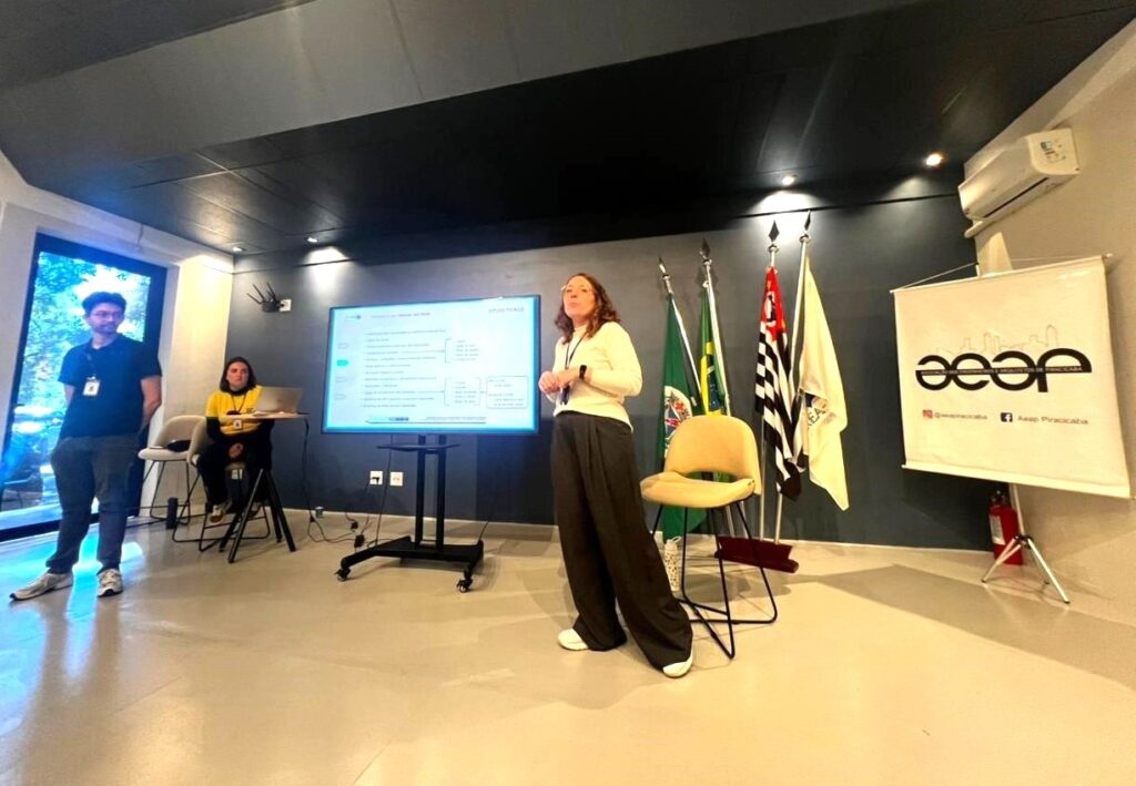 Nesta imagem, vemos uma apresentação acontecendo em uma sala bem organizada e iluminada. • A palestrante: No centro, uma mulher de blusa branca e calça escura está em pé, falando ou aguardando para começar sua apresentação. Ela está ao lado de um monitor de TV grande. • O monitor: A tela exibe um gráfico ou slide de "estudo técnico" com tópicos organizados. • A equipe: À esquerda, um homem de camiseta preta está em pé, e uma mulher vestindo uma camiseta amarela está sentada em um banco alto, provavelmente dando suporte técnico ou acompanhando a reunião. • O cenário: Ao fundo, há três bandeiras hasteadas (uma delas é a do Brasil) e, à direita, um banner branco com a logomarca "AEAP" (Associação dos Engenheiros e Arquitetos de Piracicaba). O ambiente é moderno, com paredes cinzas e piso claro.