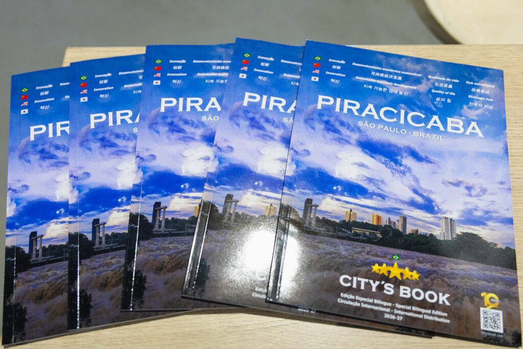 A imagem mostra quatro exemplares idênticos de um folheto ou revista dispostos em leque sobre uma superfície de madeira clara. O título principal do material é "PIRACICABA SÃO PAULO - BRAZIL", escrito em letras grandes e brancas. A capa apresenta uma fotografia panorâmica de uma paisagem natural com um rio e corredeiras em primeiro plano, e uma cidade ao fundo sob um céu azul com algumas nuvens. Na parte inferior da capa, há uma faixa com bandeiras de diversos países (Brasil, China, Estados Unidos e Coreia do Sul) acompanhadas por textos em vários idiomas, incluindo português, chinês, inglês e coreano. Esses textos destacam temas como "Inovação", "Desenvolvimento econômico sustentável" e "Qualidade de vida", enfatizando o caráter internacional e bilíngue da publicação. No canto inferior direito, está o logotipo "CITY'S BOOK" junto com a indicação de que se trata de uma edição especial bilíngue com circulação internacional para os anos de 2026-27. Também há um selo comemorativo de "10 YEARS" e um código QR, sugerindo um conteúdo interativo ou digital associado ao folheto. A iluminação da foto é clara, destacando o acabamento brilhante dos exemplares.