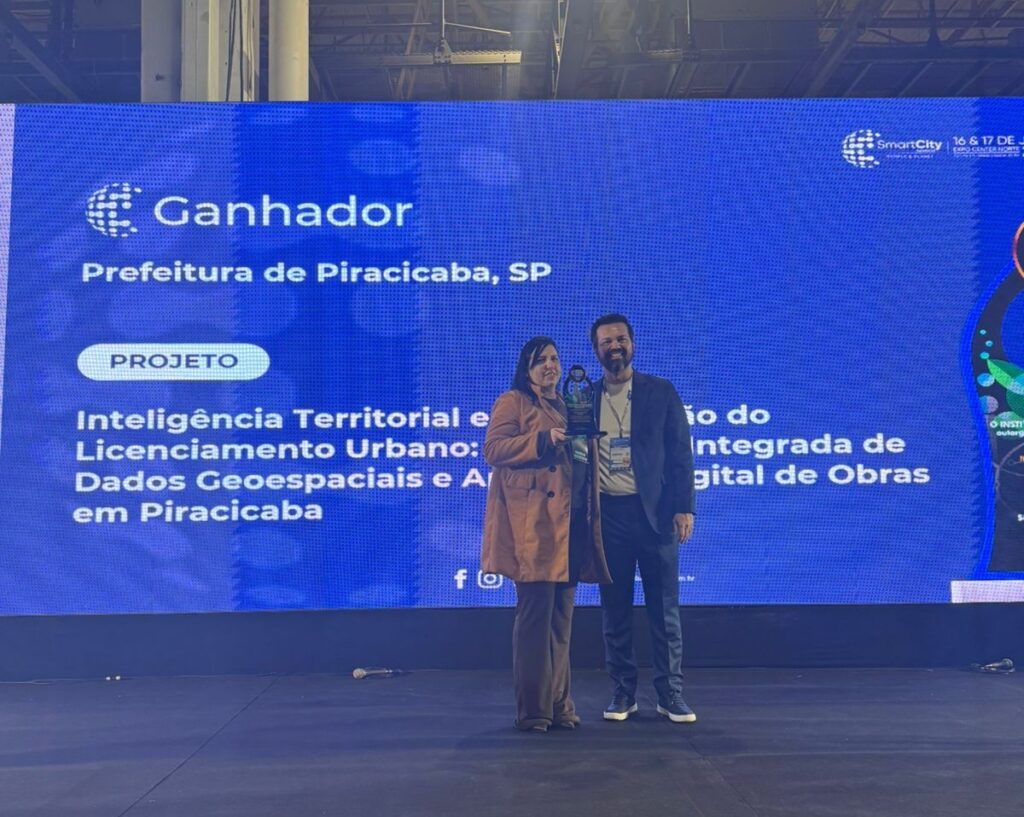 A imagem mostra uma cerimônia formal de premiação realizada em um palco. No centro da imagem, há duas pessoas posando para a foto: uma mulher à esquerda, vestindo um casaco bege claro e calças marrons, segurando um troféu, e um homem à direita, usando um blazer escuro, camisa branca e calças escuras, com um crachá pendurado no pescoço. Ambos estão sorrindo. Ao fundo, há uma grande tela digital azul com informações do evento. Nela está escrito "Ganhador", seguido pelo nome da Prefeitura de Piracicaba, SP, e o título do projeto premiado: "Inteligência Territorial e Licenciamento Urbano: Dados Geoespaciais e A... no Integrado de Digital de Obras em Piracicaba". No canto superior direito da tela, há o logotipo do evento "SmartCity". Na parte inferior da tela, aparecem ícones de redes sociais. O ambiente está iluminado por luzes de palco, e há um microfone no chão, indicando que a cerimônia provavelmente incluiu discursos ou apresentações. A imagem transmite um momento de reconhecimento e celebração de um projeto relacionado a tecnologia e gestão urbana.