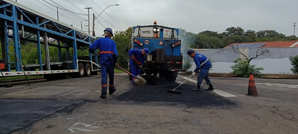 Nesta imagem, vemos três trabalhadores uniformizados realizando um serviço de manutenção asfáltica em uma rua. Eles usam uniformes azuis com faixas refletivas e capacetes de proteção. O foco central é a equipe trabalhando com um caminhão de serviço (que possui um equipamento de pavimentação, com fumaça saindo dele), espalhando massa asfáltica quente sobre a via com o auxílio de pás e rodos. À esquerda, há parte de uma carreta cegonha estacionada, e à direita, um cone de sinalização laranja sinaliza o trabalho. O cenário é ao ar livre, em um dia nublado, com vegetação e um muro ao fundo.
