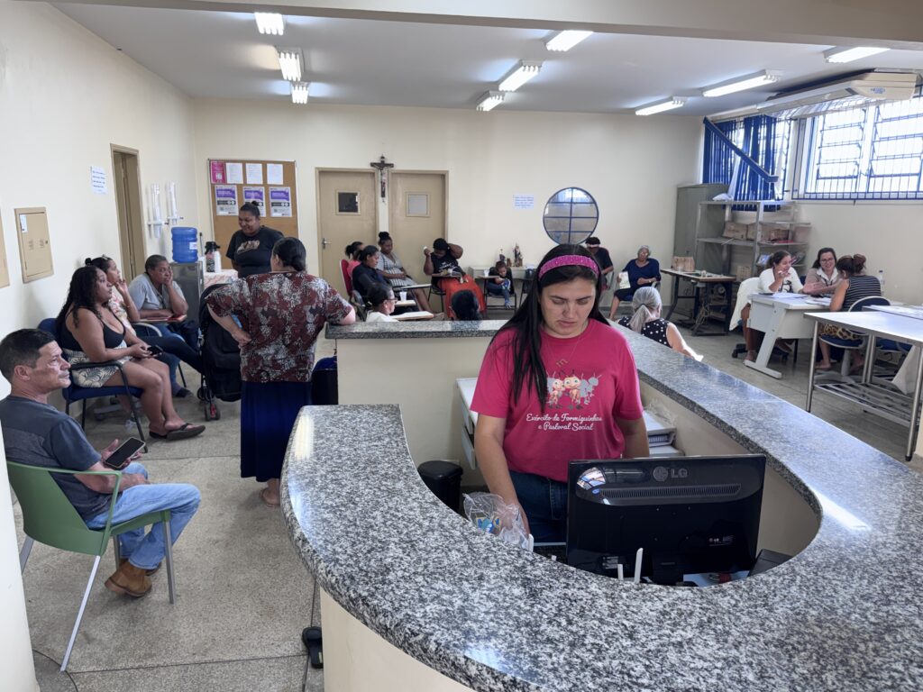 A imagem mostra o interior de um centro comunitário ou escritório de serviço social bastante movimentado. Em primeiro plano, há uma mulher vestindo uma camiseta rosa com os dizeres "Exército de Formiguinhas e Pastoral Social", que está posicionada atrás de um balcão de granito, olhando para um monitor de computador da marca LG. Ao lado dela, um homem está sentado em uma cadeira verde, olhando em direção ao centro da sala. Ao fundo, várias pessoas estão sentadas em cadeiras, aguardando ou participando de alguma atividade ou reunião. O ambiente é simples e funcional, com paredes claras, algumas portas, um quadro de avisos com papéis fixados e janelas grandes com cortinas azuis. A iluminação é feita por lâmpadas fluorescentes no teto. O local possui um clima informal e acolhedor, típico de um espaço voltado para atendimento comunitário ou ações sociais. No ambiente, também é possível ver uma bebedouro, mesas e cadeiras distribuídas pelo espaço, que tem piso de azulejo em tom neutro. A imagem é capturada a partir do balcão de atendimento, oferecendo uma visão ampla da sala e das pessoas presentes.
