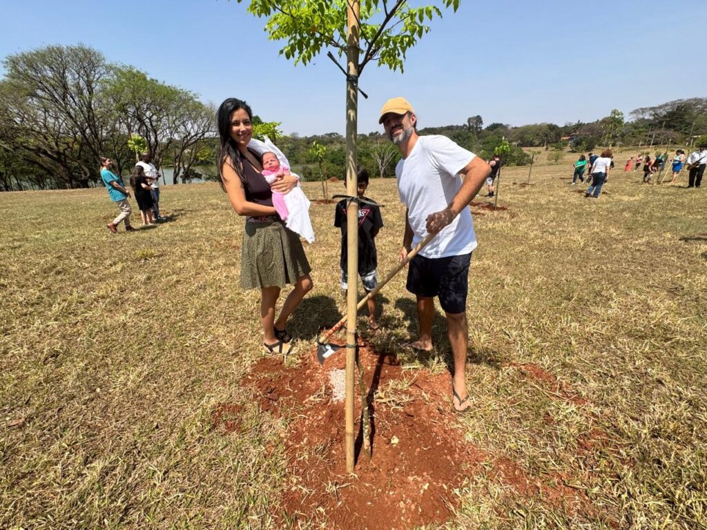 A imagem mostra uma família de quatro pessoas participando de um plantio de árvores em um campo aberto e amplo, com grama seca e áreas de terra exposta. O cenário é ensolarado, com céu azul e algumas árvores ao fundo, além de um corpo d'água visível ao longe. A mãe, vestindo uma regata preta e saia verde estampada, segura um bebê enrolado em um cobertor branco. Ao lado dela está um menino, provavelmente o filho mais velho, usando camiseta preta e shorts. O pai, de camiseta branca, boné bege e shorts escuros, está agachado ao lado de uma muda de árvore recém-plantada, segurando uma ferramenta de jardinagem para ajeitar a terra ao redor da muda. Ao fundo, outras pessoas também participam da atividade, indicando um evento comunitário de plantio de árvores. A cena transmite um momento de união familiar e cuidado com o meio ambiente.