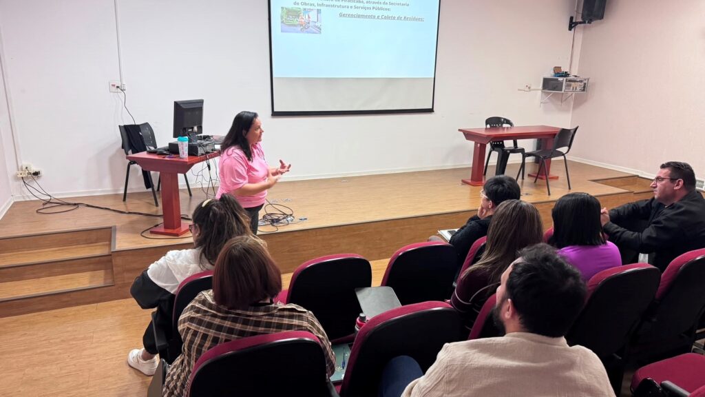 A imagem mostra outro momento da mesma palestra sobre gestão de resíduos em Piracicaba.Cena:Palestrante: A mulher de camiseta rosa agora está no meio do palco, gesticulando enquanto fala. Ela saiu de trás da mesa e interage diretamente com o público.Slide projetado: Continua sobre "Gerenciamento e Coleta de Resíduos" da Prefeitura de Piracicaba, via Secretaria de Obras, Infraestrutura e Serviços Públicos. Dá pra ver uma foto pequena de um caminhão de coleta.Público: As mesmas pessoas das fotos anteriores estão sentadas nas poltronas vermelhas, prestando atenção. Alguns estão com caderno ou prancheta no colo. O pessoal de xadrez, roxo e jaqueta bege aparece de novo.Ambiente: Auditório com palco de madeira e dois degraus. Tem uma mesa vermelha com computador à esquerda e outra mesa vazia à direita. Paredes brancas, caixa de som e tomadas aparentes.É um momento mais dinâmico da apresentação, com a palestrante explicando o conteúdo de forma mais próxima dos participantes. Faz parte da mesma sequência de capacitação sobre os deveres da Prefeitura na coleta de resíduos.