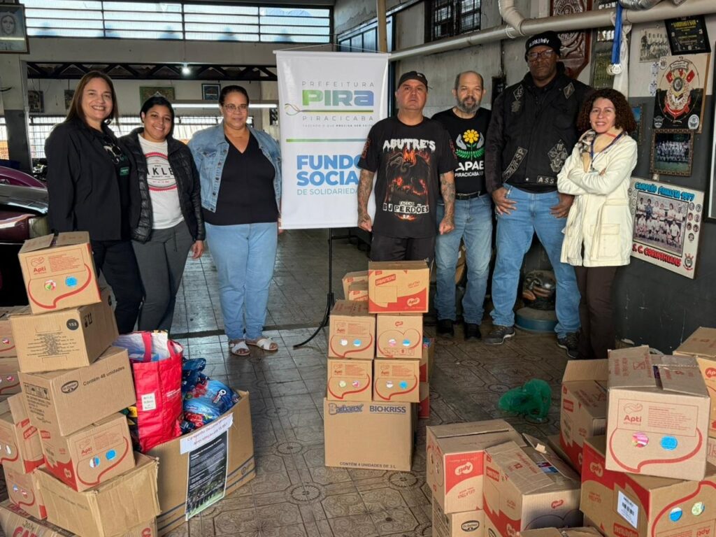 A imagem registra um momento de entrega de doações, possivelmente alimentos, com a participação de um grupo de pessoas e representantes do Fundo Social de Solidariedade da Prefeitura de Piracicaba. * O grupo, composto por cinco homens e três mulheres, está posicionado atrás de uma grande quantidade de caixas de papelão empilhadas, que contêm produtos da marca Apti e itens diversos. * Ao centro, destaca-se um banner branco com o logotipo da Prefeitura de Piracicaba e a inscrição Fundo Social de Solidariedade. * Os participantes vestem trajes casuais e uniformes de organizações, incluindo um homem com uma camiseta do motoclube Abutre's e outro com uma camiseta da APAE Piracicaba. * O cenário parece ser o interior de um galpão ou sede social, com piso de cerâmica, paredes decoradas com quadros e itens esportivos, incluindo uma moldura com uma foto de um time do S.C. Corinthians. * À esquerda, há uma caixa de papelão com um cartaz informativo afixado, indicando uma campanha de arrecadação. O ambiente é iluminado por luz artificial e natural.