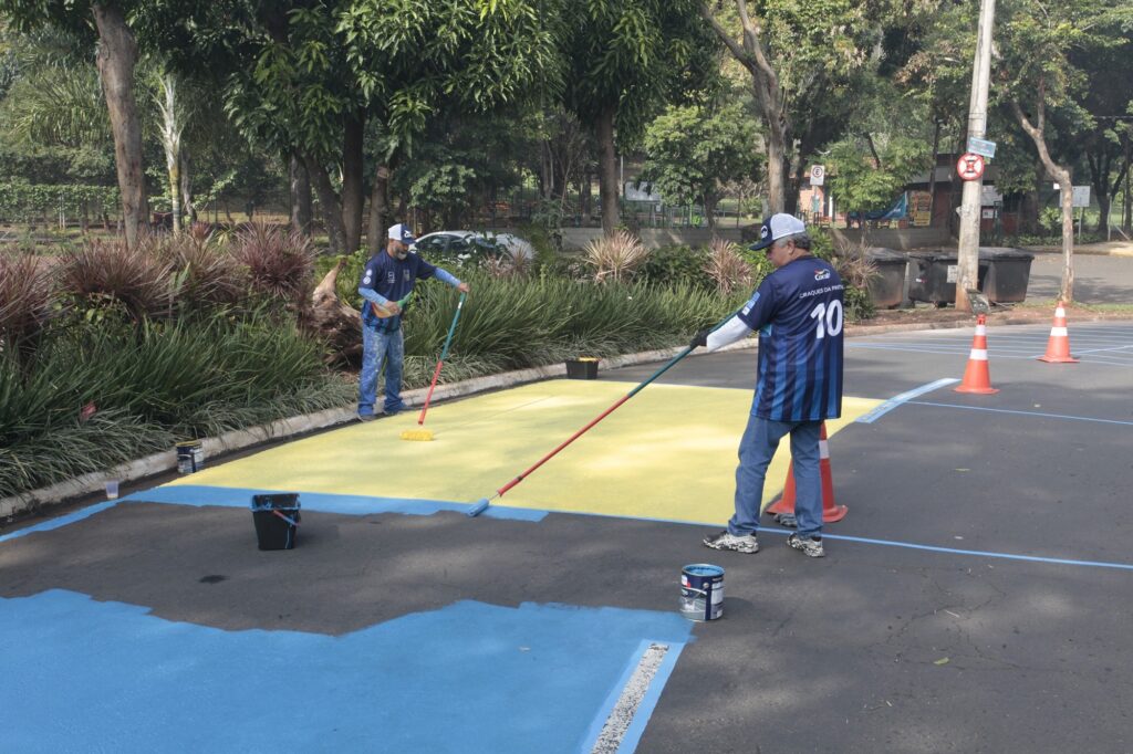 A imagem mostra dois trabalhadores pintando uma área de estacionamento ao ar livre. Ambos vestem camisetas esportivas azuis com detalhes em azul-marinho, com a marca "Coral" e a frase "CRAQUES DA PINTURA" estampadas nas costas, além do número 10 em uma delas. Eles estão usando bonés e calças jeans. Os homens estão aplicando tinta amarela na superfície do asfalto usando rolos de pintura com cabos longos. A área já tem marcações azuis, possivelmente delimitando os espaços a serem pintados. Ao redor, há latas de tinta, cones de sinalização e um carro branco estacionado. Ao fundo, é possível ver árvores e vegetação, indicando que o local é um espaço aberto, possivelmente um parque ou uma área pública. A cena ocorre durante o dia, com luz natural abundante.