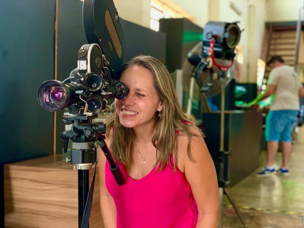 A imagem mostra uma mulher sorridente posicionada atrás de uma câmera de cinema profissional antiga. Ela veste uma blusa regata na cor rosa vibrante e um colar delicado. A câmera, montada sobre um tripé, possui uma torre de lentes giratória e um chassi de filme na parte superior, remetendo a equipamentos clássicos de filmagem. Ao fundo, em um ambiente que parece ser um estúdio ou set de filmagem, um homem aparece desfocado, vestindo uma camiseta branca e bermuda azul, trabalhando próximo a um refletor de iluminação profissional montado em um pedestal. O cenário é iluminado artificialmente, sugerindo um ambiente de produção audiovisual.